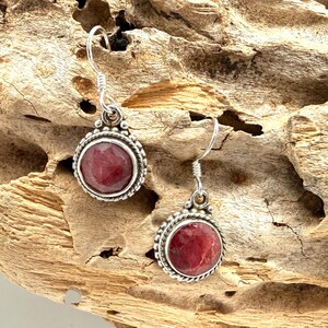 May include: A pair of silver earrings with a round, deep red gemstone center. The earrings have a beaded border and a simple hook closure. The background is a piece of weathered wood.