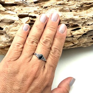 K&ouml;nnte beinhalten: Ein silberner Ring mit einem ovalen, schwarzen Edelstein wird auf einer Hand gezeigt. Der Ring hat ein verziertes Design um den Stein herum. Der Hintergrund umfasst ein St&uuml;ck Treibholz.