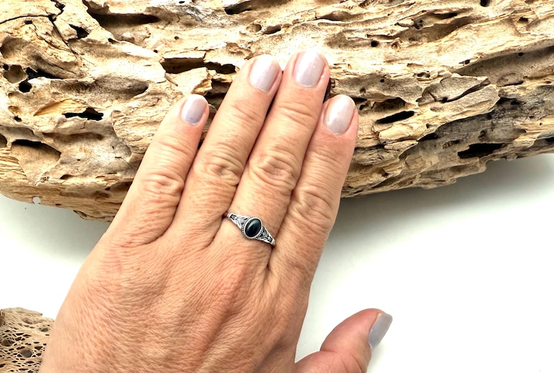 K&ouml;nnte beinhalten: Ein silberner Ring mit einem ovalen, schwarzen Edelstein wird auf einer Hand gezeigt. Der Ring hat ein verziertes Design um den Stein herum. Der Hintergrund umfasst ein St&uuml;ck Treibholz.