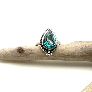 K&ouml;nnte beinhalten: Ein silberner Ring mit einem tropfenf&ouml;rmigen Einlegeteil aus Abalone-Muschel. Der Ring hat ein Perlenmuster um den Rand der Fassung.