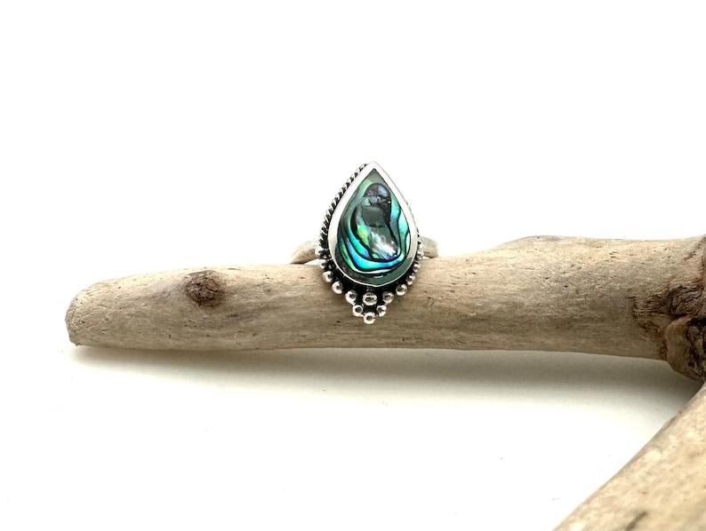 K&ouml;nnte beinhalten: Ein silberner Ring mit einem tropfenf&ouml;rmigen Einlegeteil aus Abalone-Muschel. Der Ring hat ein Perlenmuster um den Rand der Fassung.