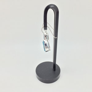 Puede incluir: Un pendiente de plata con un colgante de piedra azul que cuelga de un soporte de pendientes negro.