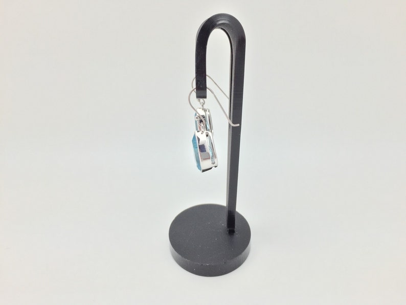 Puede incluir: Un pendiente de plata con un colgante de piedra azul que cuelga de un soporte de pendientes negro.