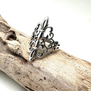 May include: A silver ring with a delicate, intricate design featuring multiple fleur-de-lis motifs. The ring is set on a piece of driftwood.