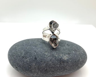 Può includere: Un anello d'argento con due cristalli di quarzo fumé grezzi. I cristalli sono incastonati in un design geometrico, con una fascia che avvolge il dito. L'anello è esposto su una pietra grigia su sfondo bianco.