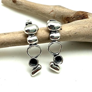 May include: A pair of silver-toned earrings featuring a vertical arrangement of smooth, rounded pebble-like shapes and an open circular link. The earrings have a polished, reflective surface and a drop design that hangs from a post-style backing.