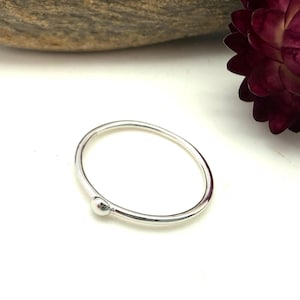 May include: A thin, polished silver ring featuring a single small, smooth spherical bead accent. The band is circular and maintains a consistent, slender thickness throughout its design, showcasing a reflective metallic finish on its surface.