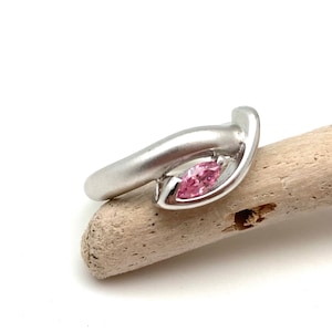 May include: A silver ring featuring a pink marquise-shaped gemstone set in the centre. The ring has a curved band that wraps around the finger.