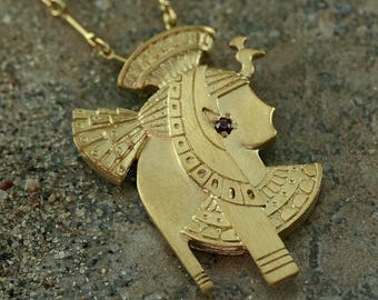 May include: Gold-toned pendant necklace featuring a profile of an Egyptian figure. The pendant has intricate details, including a headdress and a small, dark red gemstone. The necklace is displayed on a textured, neutral-toned surface.