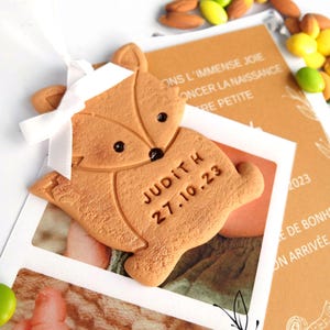 May include: A light brown fox-shaped ornament with the name "Judith" and the date "27.10.23" engraved on it. The fox has black eyes and a white ribbon. The ornament is placed over a photo and a card with French text and candies.