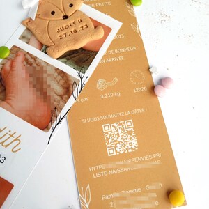 May include: A baby announcement card with a fox-shaped cookie and a photo. The card is tan with text in French, including the baby's name, Judith, and birthdate, 27.10.23. The card also includes the baby's weight, 3.210 kg, and length, 8 cm.
