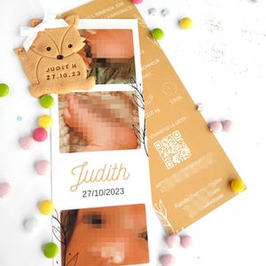 May include: A baby announcement card with two photos and the name "Judith" and date "27/10/2023". A fox-shaped cookie with the name "Judith H" and date "27.10.23" is attached. Colorful candies and a brown card are scattered around.