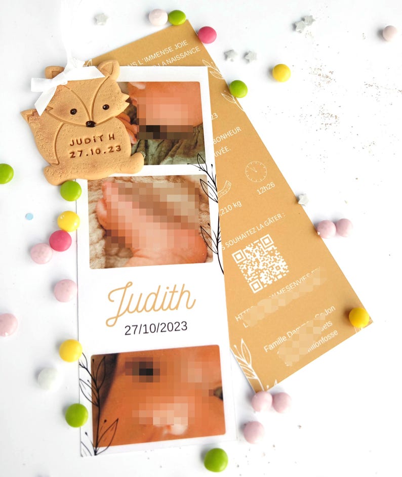 May include: A baby announcement card with two photos and the name "Judith" and date "27/10/2023". A fox-shaped cookie with the name "Judith H" and date "27.10.23" is attached. Colorful candies and a brown card are scattered around.