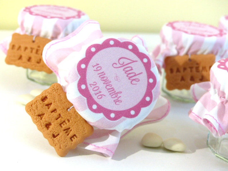 May include: Pink and white striped fabric jars with a pink circle label that says "Jade" and "19 novembre 2016". Each jar has a small, round, brown cookie with the text "BAPT&Ecirc;ME A&J" on it.