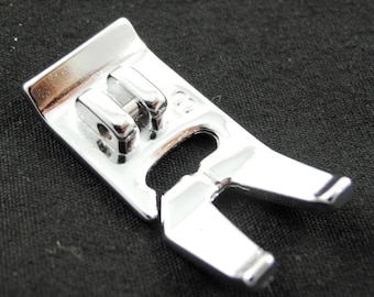 May include: A silver sewing machine foot with a small metal plate on the top. The foot has a curved shape with two prongs on the bottom.