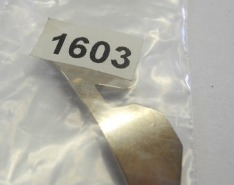 May include: A silver metal part with a number "1603" printed on a white label. The part is shaped like a hook with a small hole at the bottom.