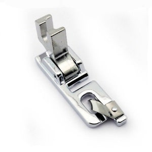 May include: A silver sewing machine foot with a metal guide and a spring-loaded mechanism.