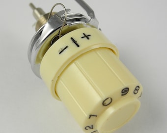 May include: Close-up of a sewing machine tension dial. The dial is off-white with black markings, including numbers and plus/minus symbols. A silver metal component and a wire loop are also visible.