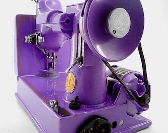 May include: A vintage Singer sewing machine finished in a vibrant purple enamel paint. The machine features a prominent handwheel, a black drive belt, a metallic gold circular emblem, and a black electrical power port located on the base.