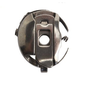 May include: A silver-colored sewing machine bobbin case. The metal case has a circular shape with a central opening and a hinged latch. A curved metal piece extends from the side. The bobbin case is designed to hold the bobbin and thread for sewing.