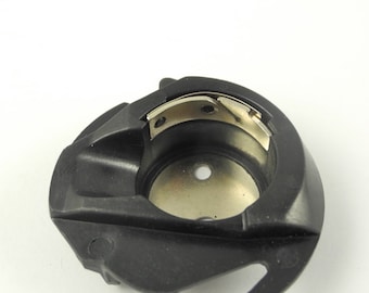 May include: Black plastic bobbin case for a sewing machine. The bobbin case has a metal hook and a metal bobbin holder.