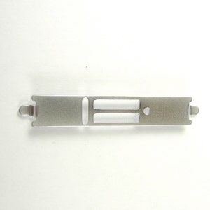 May include: A silver-colored metal computer expansion slot cover featuring a rectangular shape with rounded ends, two horizontal rectangular cutouts, and a small circular hole on the right side for mounting purposes.