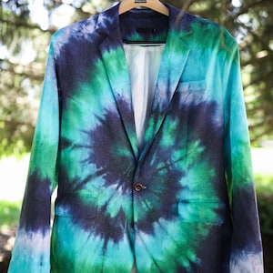 May include: A tie-dye blazer in shades of blue and green hangs from a wooden hanger. The jacket features a classic two-button closure and a white interior lining. The blazer is displayed outdoors, with a blurred background of trees.