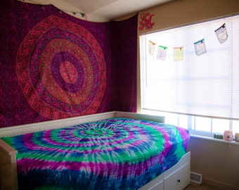 Può includere: Camera da letto con struttura letto bianca e copriletto tie-dye blu, verde e viola. Un grande arazzo con un disegno mandala circolare è appeso sopra il letto. La stanza ha una finestra con persiane e foto decorative.