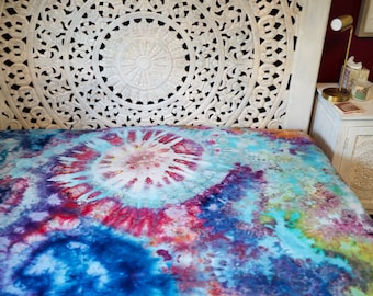 Può includere: Un copriletto tie-dye colorato con un motivo a mandala bianco al centro. Il copriletto è su un letto con una testata bianca con un motivo a mandala intagliato.