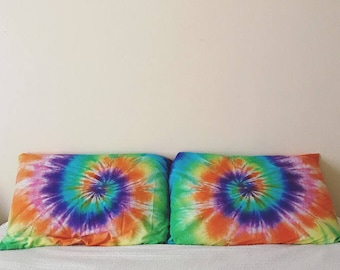 Può includere: Due federe per cuscini in tie-dye con un motivo a spirale in tonalità di arancione, giallo, verde, blu e viola. Le federe sono su un letto bianco.