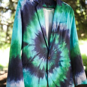 May include: A tie-dye blazer in shades of teal, green, and navy blue hangs from a wooden hanger. The jacket features a classic two-button closure, a notched lapel, and two front pockets. The interior lining is a light color.