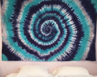 May include: A large, rectangular tapestry with a tie-dye spiral design. The pattern features concentric circles in shades of teal, navy blue, gray, and white. The tapestry is hanging above a bed with white pillows and a white sheet.