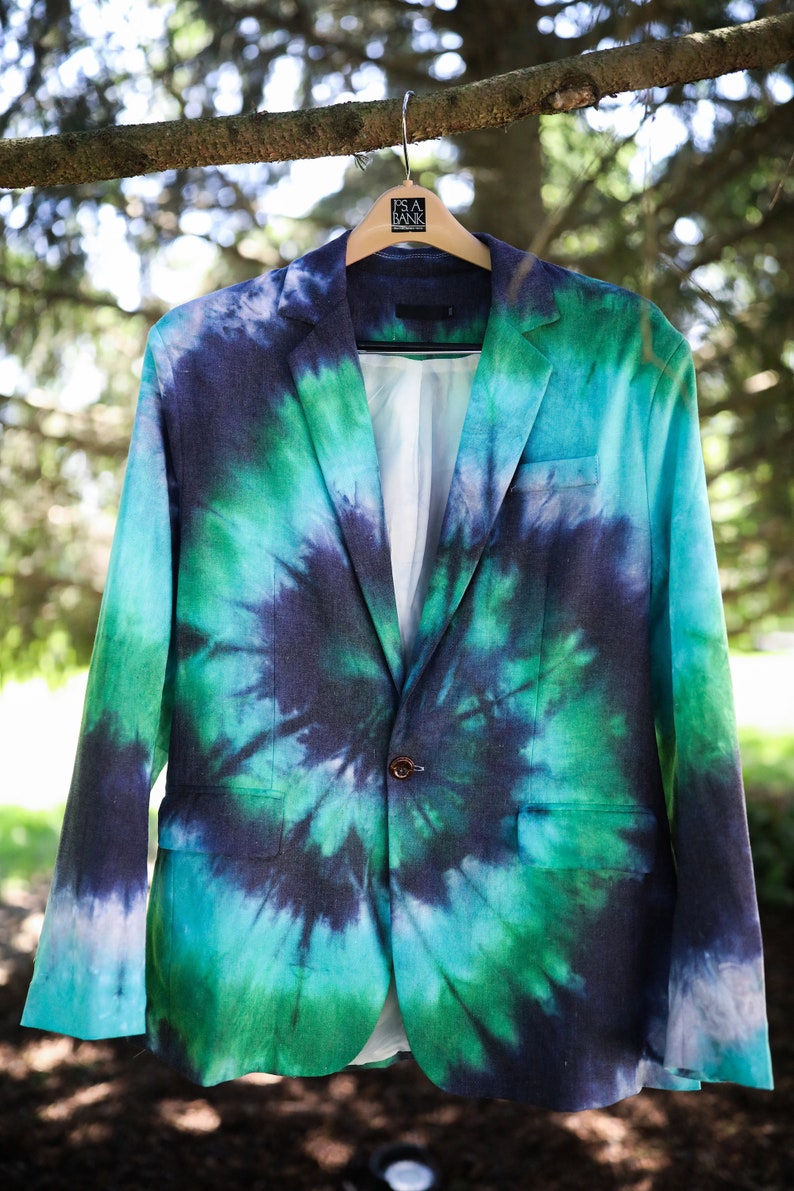 May include: A tie-dye blazer in shades of blue and green hangs from a wooden hanger. The jacket features a classic two-button closure and a white interior lining. The blazer is displayed outdoors, with a blurred background of trees.