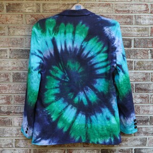 May include: A blazer with a vibrant tie-dye pattern in shades of blue, green, and purple. The jacket is hanging on a wooden hanger against a brick wall. The tie-dye design creates a spiral effect.