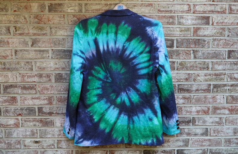 May include: A blazer with a vibrant tie-dye pattern in shades of blue, green, and purple. The jacket is hanging on a wooden hanger against a brick wall. The tie-dye design creates a spiral effect.