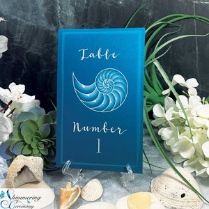 May include: Blue table number card with a white nautilus shell design and the text "Table Number 1".
