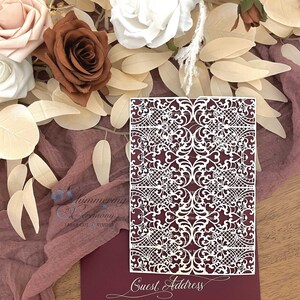 May include: A laser-cut invitation with intricate white filigree design over a deep burgundy background. The words "Guest Address" are scripted at the bottom. The invitation is surrounded by flowers and fabric.