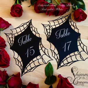 Könnte beinhalten: Zwei schwarze Tischnummernkarten mit einem Spinnennetz-Design. Die Karten liegen auf einer Holzoberfläche mit roten Rosen. Der Text auf den Karten lautet "Tisch 15" und "Tisch 17".