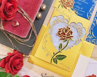 May include: A collection of wedding invitations and accessories. The invitations feature laser-cut rose designs in gold, with blue and white accents. A red rose, jewelry, and a book are also present. The text "Shimmering Ceremony Laser Cut Invitations" is visible.
