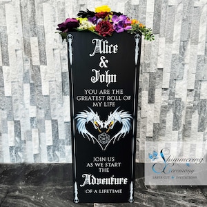 May include: Black wedding sign with white text that reads "Alice & John You are the greatest roll of my life Join us as we start the Adventure of a lifetime". The sign features a white dragon design with a 20-sided die in the center.