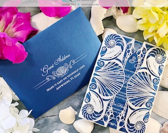 May include: A white laser cut wedding invitation with a seashell design. The invitation is folded in half and has a blue envelope with the text "Guest Address" and "Make it easy on yourself lane, anywhere, FL 33436".