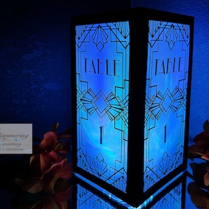 Puede incluir: Un porta números de mesa negro con un diseño geométrico y una luz azul que brilla a través. El número "1" es visible en el centro del porta números. El texto "TABLE" es visible en la parte superior del porta números.