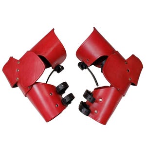 Pode incluir: Par de braçadeiras de couro vermelho com tiras e fivelas pretas. As braçadeiras têm um design curvo e são rebitadas. Adequadas para fantasias ou reconstituições históricas.
