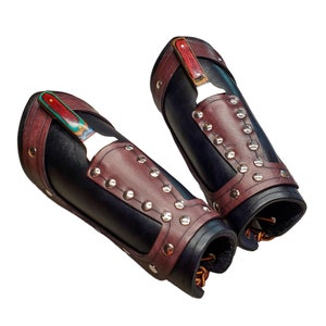 May include: A pair of black leather arm guards with brown leather accents and silver studs. Each guard has a small, colorful wooden piece attached to the top.