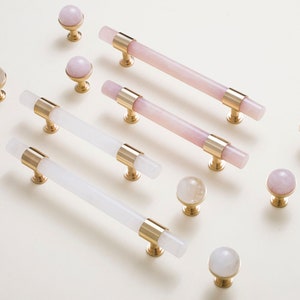 May include: Assortment of pink and white marble cabinet knobs and pulls with gold accents. The knobs are round and the pulls are long and slender.