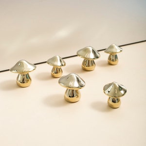 Może przedstawiać: Zestaw sześciu złotych ceramicznych figurek grzybów w różnych rozmiarach. Grzyby są wszystkie w błyszczącym wykończeniu i mają gładki, zaokrąglony kształt.