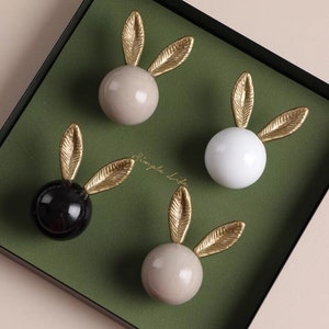 May include: Four ceramic bunny-shaped knobs with gold leaf ears. The knobs are in shades of white, black, and beige. The knobs are displayed on a green surface.