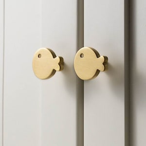 Könnte beinhalten: Zwei goldfarbene, fischförmige Schrankknöpfe mit schlichtem Design.