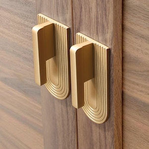 Op de afbeelding: Twee gouden kastdeurgrepen met een decoratief, geometrisch ontwerp. De grepen zijn gemonteerd op een houten kastdeur.
