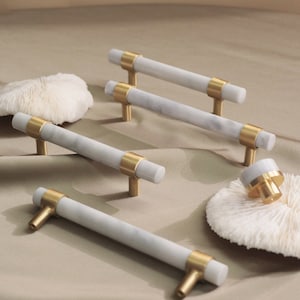 May include: Five white marble cabinet knobs with gold accents. The knobs are arranged on a beige surface.